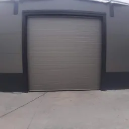 Zamknięta, segmentowa brama garażowa w kolorze szarym, zamontowana w szarej elewacji budynku przemysłowego, widok z poziomu posadzki betonowej.