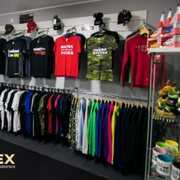 Wnętrze sklepu odzieżowego w Białymstoku z szerokim wyborem koszulek z nadrukami patriotycznymi i humorystycznymi, oraz odzieży w różnych kolorach, eksponowane na wieszakach i półkach.