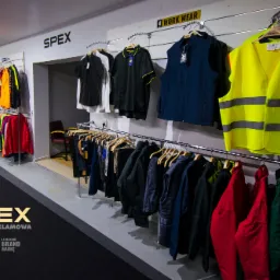 Ekspozycja odzieży roboczej i sportowej w sklepie, widoczne wieszaki z r&oacute;żnorodnymi ubraniami, w tym kamizelki odblaskowe, koszulki polo i kurtki, na ścianie logo firmy SPEX