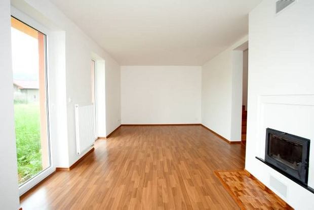 Puste, jasne wnętrze z drewnianą podłogą, białymi ścianami i wnęką kominkową z czarnym wkładem, widok na zieleń przez okno.