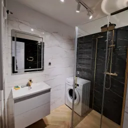 Nowoczesna łazienka z kabiną prysznicową typu walk-in, marmurowymi płytkami, umywalką z szafką i pralką. Oświetlenie punktowe. Parkiet ułożony w jodełkę.