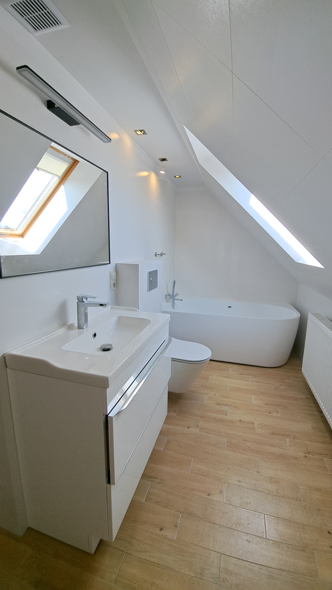 Jasna łazienka na poddaszu z oknem dachowym. Umywalka z szafką, toaleta, wanna i grzejnik. Płytki podłogowe imitujące drewno. Minimalistyczny styl.