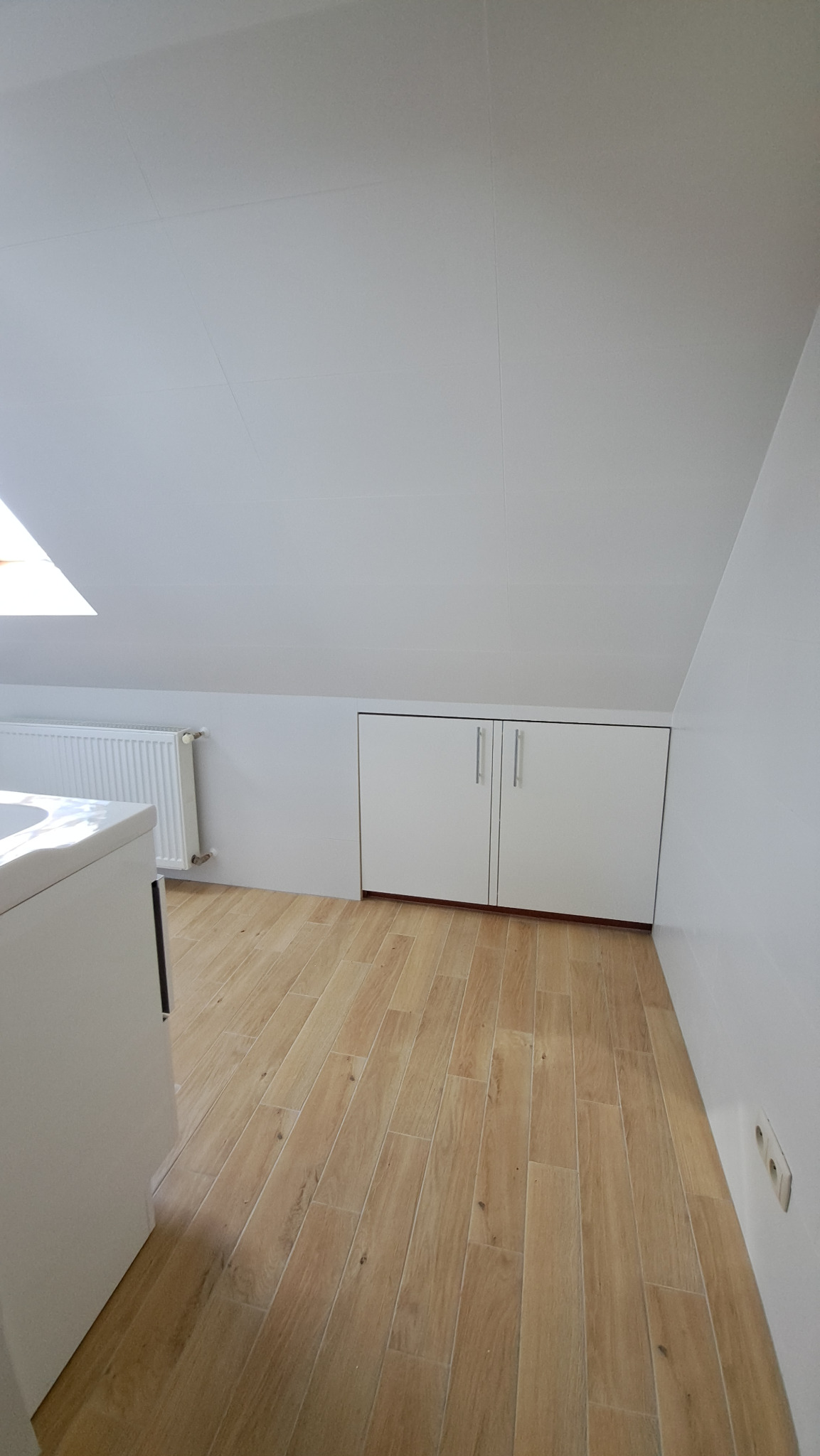 Jasne wnętrze poddasza w Sławnie z drewnopodobną podłogą, białymi szafkami w zabudowie i grzejnikiem pod oknem dachowym. Minimalistyczny styl wykończenia.