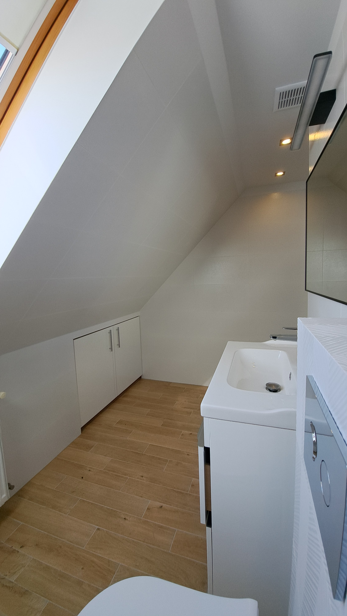 Jasna łazienka na poddaszu ze skosami, wykończona w bieli i drewnie. Umywalka z szafką, drewnopodobne płytki na podłodze, zabudowa pod skosem. Minimalistyczny styl.