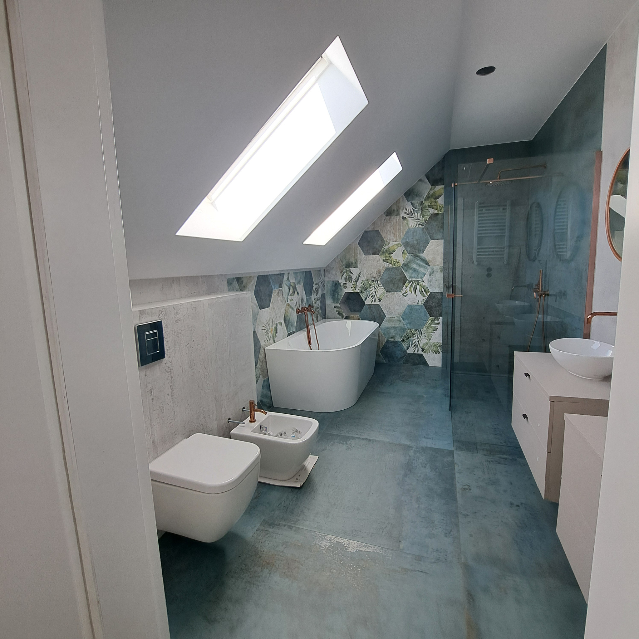 Nowoczesna łazienka na poddaszu z wanną wolnostojącą, toaletą, bidetem i kabiną prysznicową. Uwagę przykuwają heksagonalne płytki z motywem roślinnym i okna dachowe.