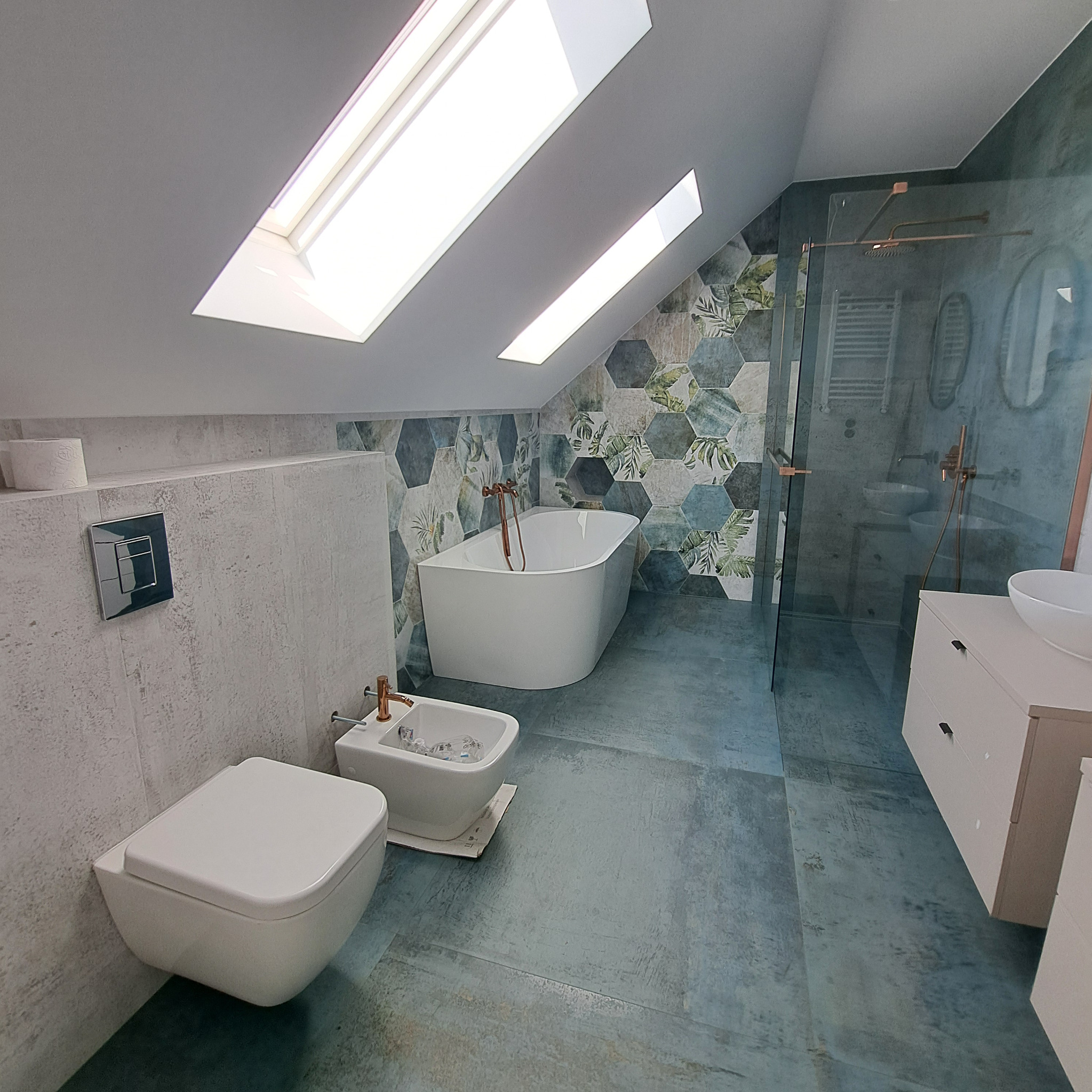 Kompletna łazienka na poddaszu z oknami dachowymi. Uwagę przykuwają heksagonalne płytki z motywem roślinnym na ścianie za wanną i w kabinie prysznicowej.