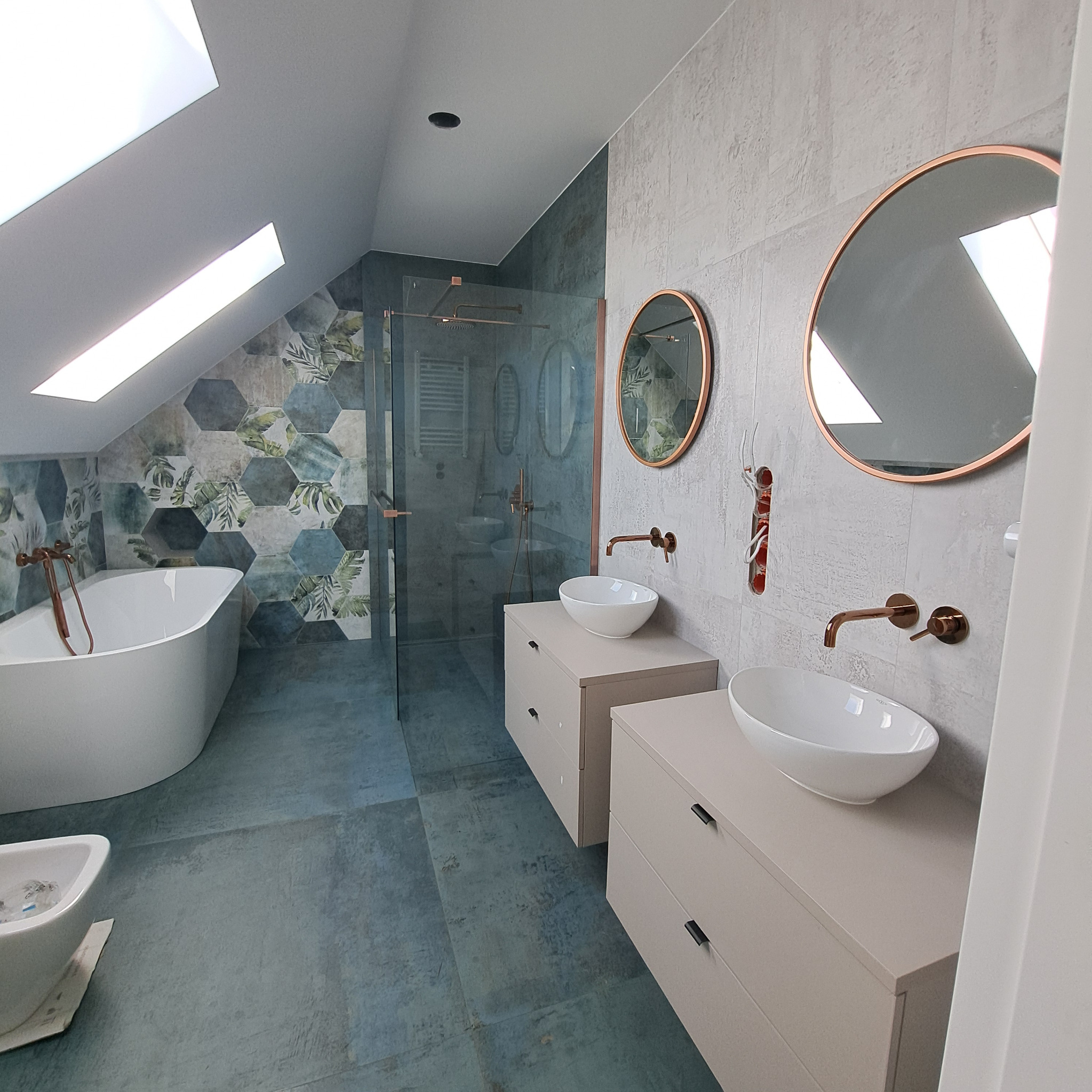 Stylowa łazienka na poddaszu z wanną, kabiną prysznicową i umywalkami na szafkach w kolorze beżowym. Okrągłe lustra z miedzianą ramą i armatura w odcieniu miedzi.