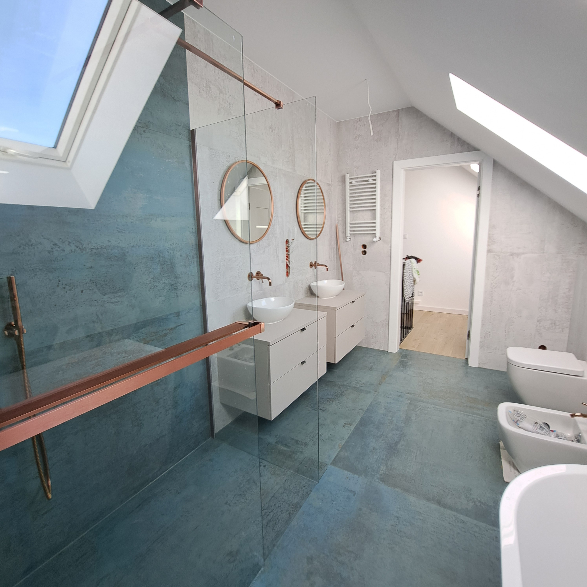 Nowoczesna łazienka na poddaszu z kabiną prysznicową, umywalkami na szafkach i oknami dachowymi. Dominuje niebiesko-szara kolorystyka z akcentami miedzianymi.