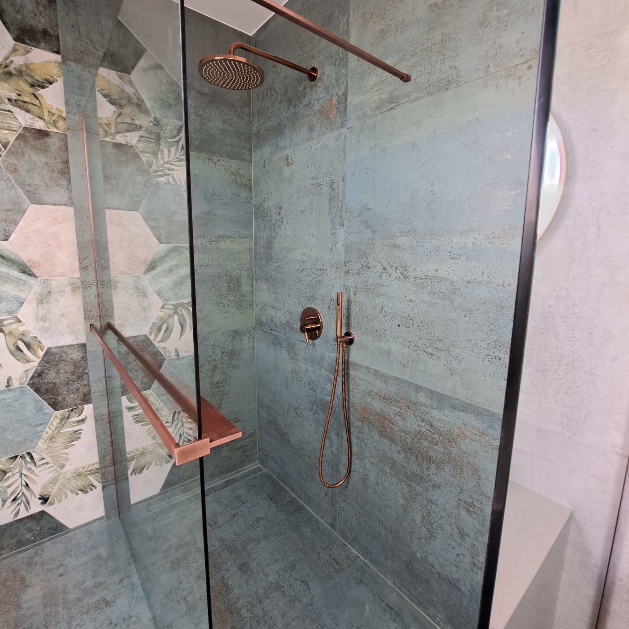 Nowoczesna kabina prysznicowa z miedzianą armaturą i heksagonalnymi płytkami z motywem liści, oddzielona szybą. Minimalistyczny design.