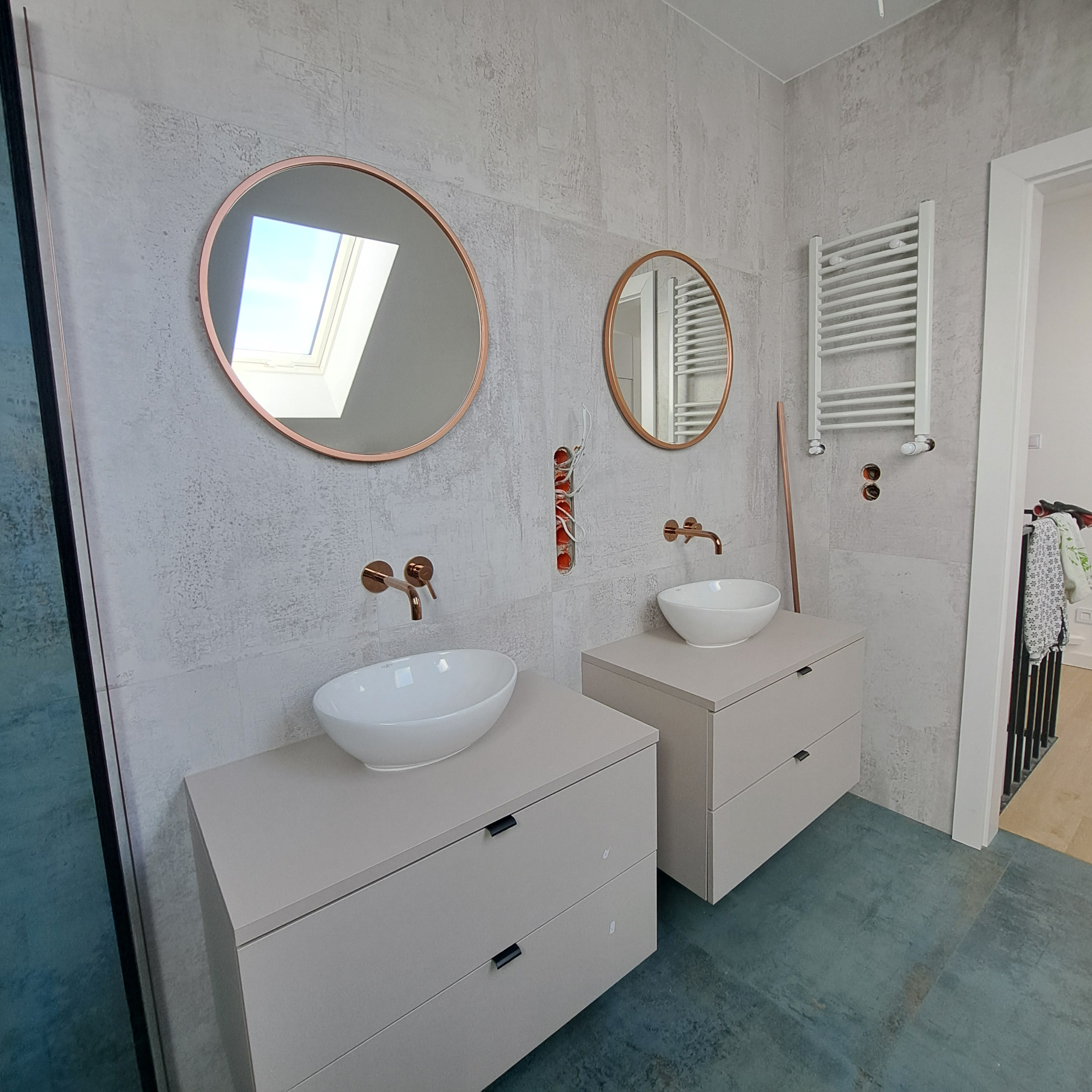 Wykończona łazienka z dwoma umywalkami nablatowymi, okrągłymi lustrami w miedzianych ramach i grzejnikiem drabinkowym. Widoczne fragmenty instalacji wodnej w ścianie.