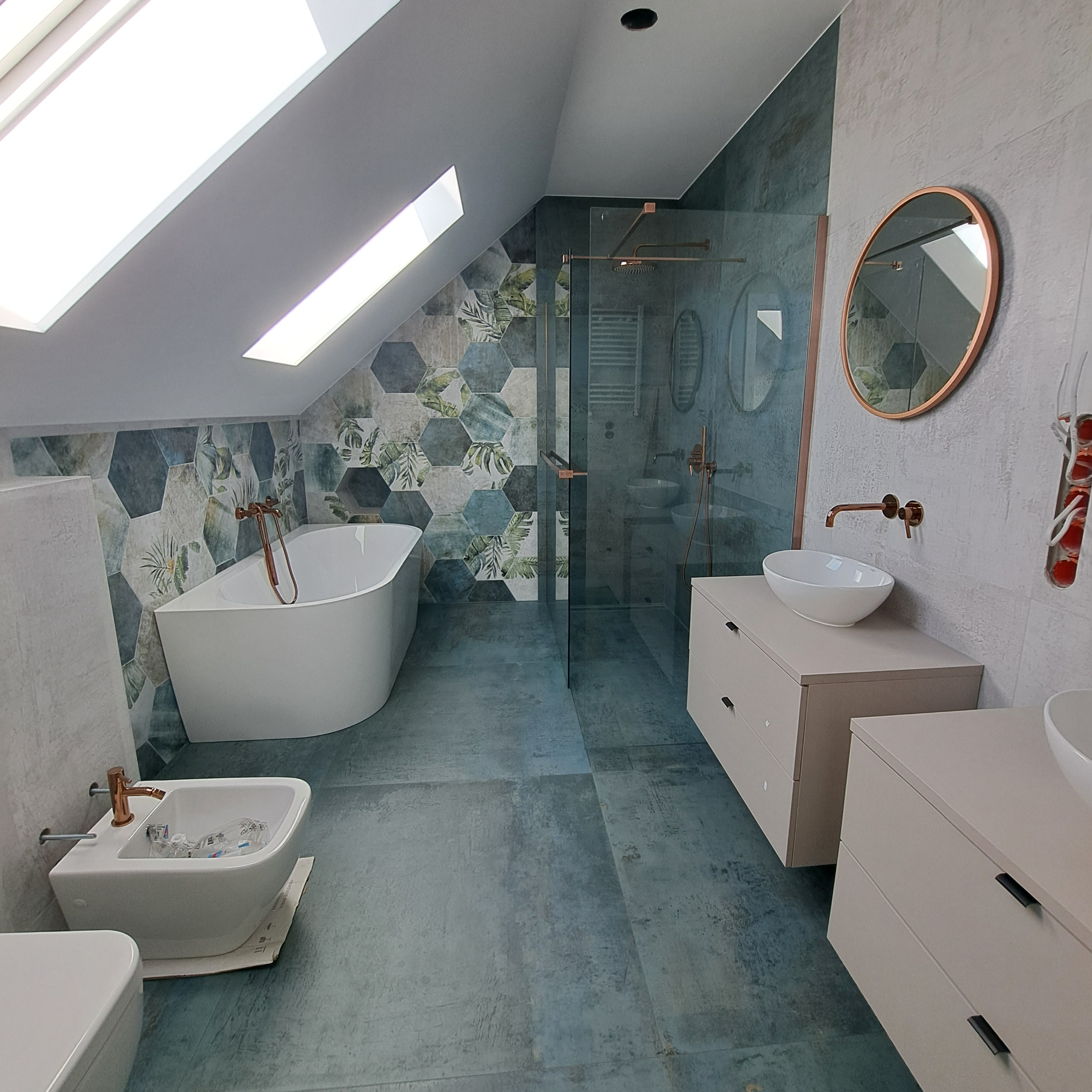 Nowoczesna łazienka na poddaszu z wanną, bidetem i szafkami w odcieniach beżu. Uwagę zwracają heksagonalne płytki z motywem roślinnym i okrągłe lustro z miedzianą ramą.