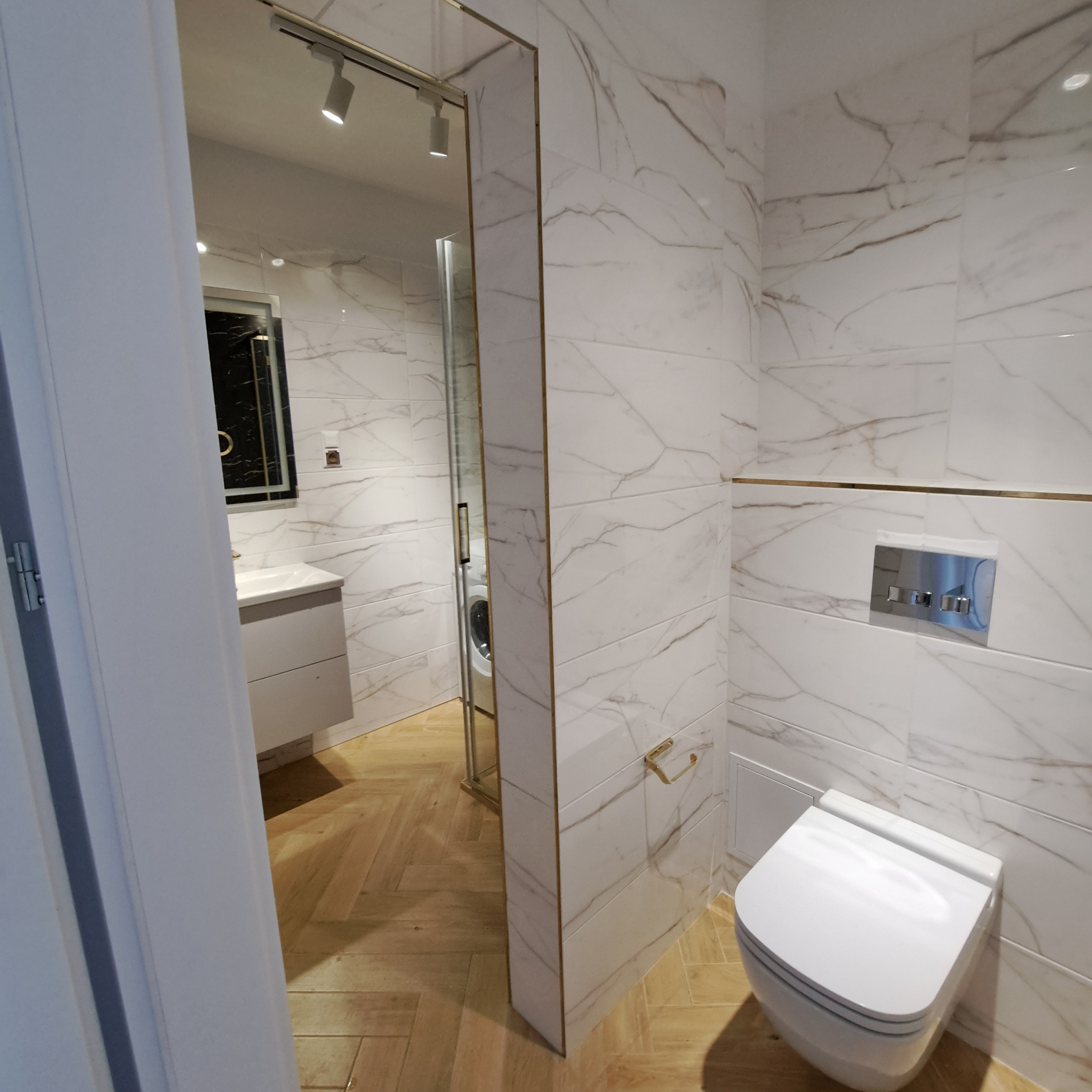 Elegancka łazienka w bieli i złocie: marmurowe płytki, drewniana podłoga w jodełkę, toaleta podtynkowa. Widok na umywalkę i pralkę przez szklaną kabinę.