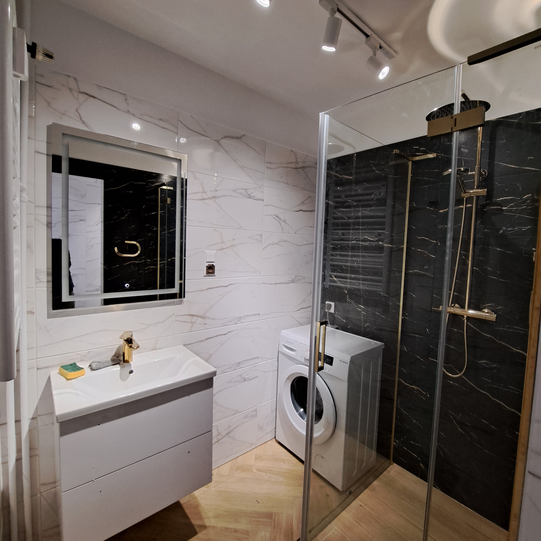 Nowoczesna łazienka z kabiną prysznicową typu walk-in, marmurowymi płytkami, umywalką z szafką i pralką. Oświetlenie punktowe. Parkiet ułożony w jodełkę.