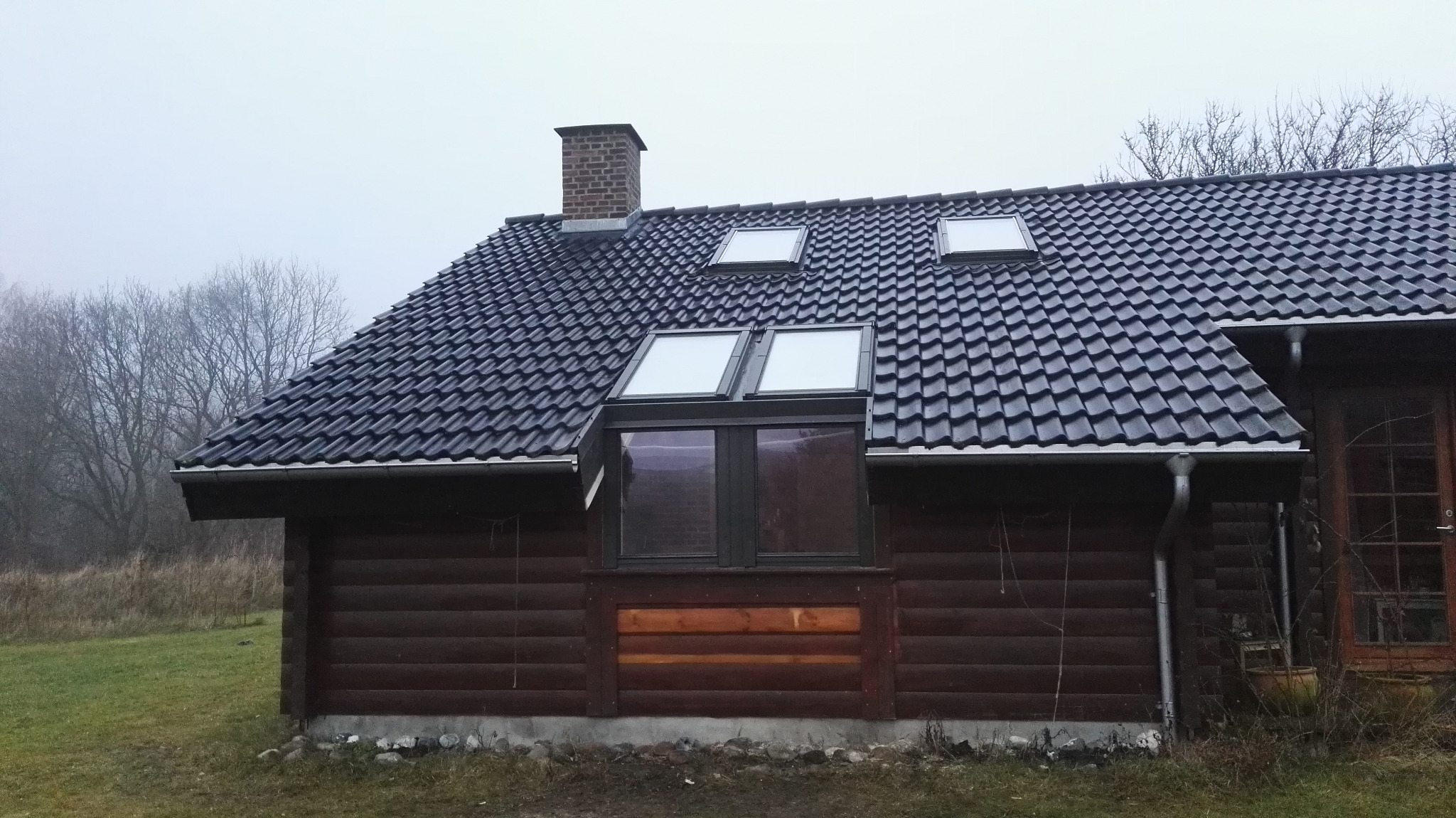 Dom z ciemnego drewna z czarnym dachem z dachówki ceramicznej i oknami dachowymi, komin z cegły, widok z zewnątrz w pochmurny dzień.