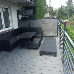 Zadaszony balkon z szarym deckingiem, rattanowym zestawem mebli ogrodowych w kolorze czarnym i metalową balustradą, z widokiem na zielony ogród.
