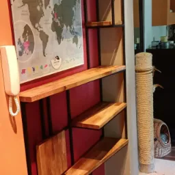 Metalowy regał z drewnianymi półkami na tle czerwonej ściany z mapą świata i beżowej ściany z drapakiem dla kota.