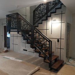 M2M SPÓŁKA Z OGRANICZONĄ ODPOWIEDZIALNOŚCIĄ - Nowoczesne schody drewniane z metalową balustradą o geometrycznym wzorze, w trakcie montażu, z widocznymi materiałami zabezpieczającymi podłogę.
