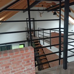 M2M SPÓŁKA Z OGRANICZONĄ ODPOWIEDZIALNOŚCIĄ - Wnętrze domu z antresolą i metalową balustradą, widok z góry na schody z drewnianymi stopniami, dekoracja w postaci czaszki zwierzęcia na ścianie pod skośnym dachem z drewnianą konstrukcją.
