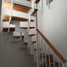 Nowoczesne schody z drewnianymi stopniami i białą metalową konstrukcją, mocowane śrubami, z białą balustradą, w jasnym wnętrzu.