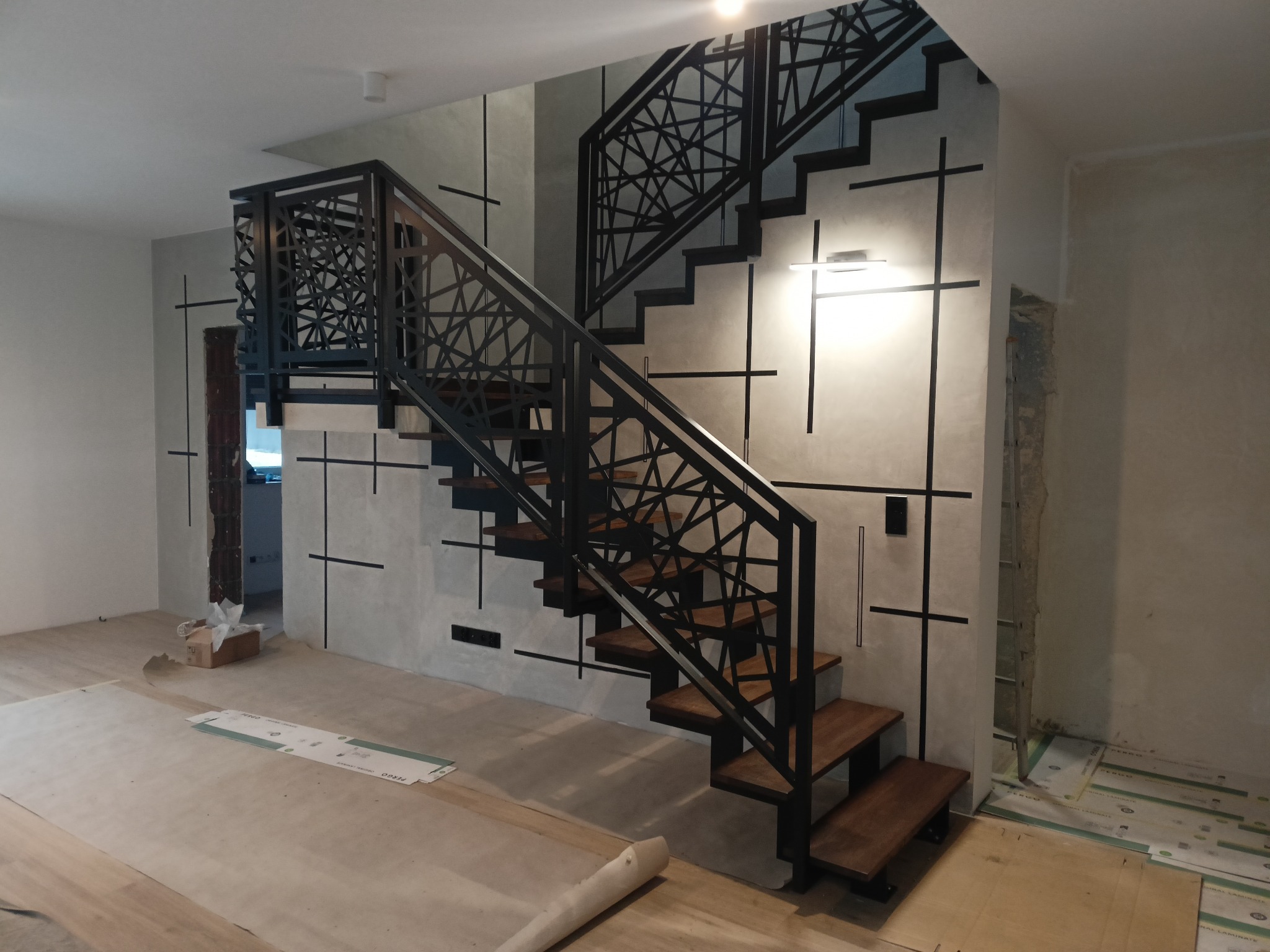 Nowoczesne schody drewniane z metalową balustradą o geometrycznym wzorze, w trakcie montażu, z widocznymi materiałami zabezpieczającymi podłogę.