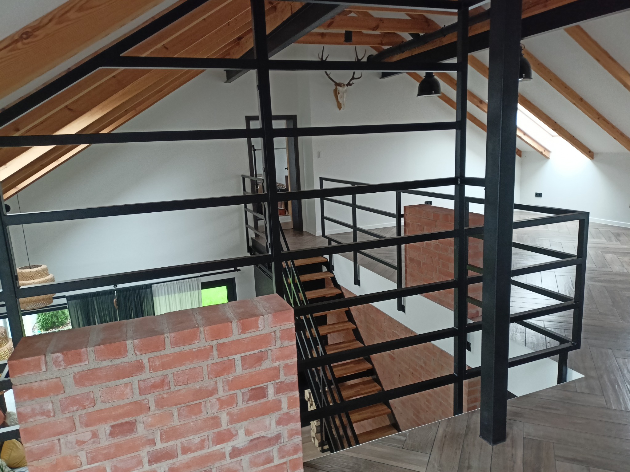 Wnętrze domu z antresolą i metalową balustradą, widok z góry na schody z drewnianymi stopniami, dekoracja w postaci czaszki zwierzęcia na ścianie pod skośnym dachem z drewnianą konstrukcją.