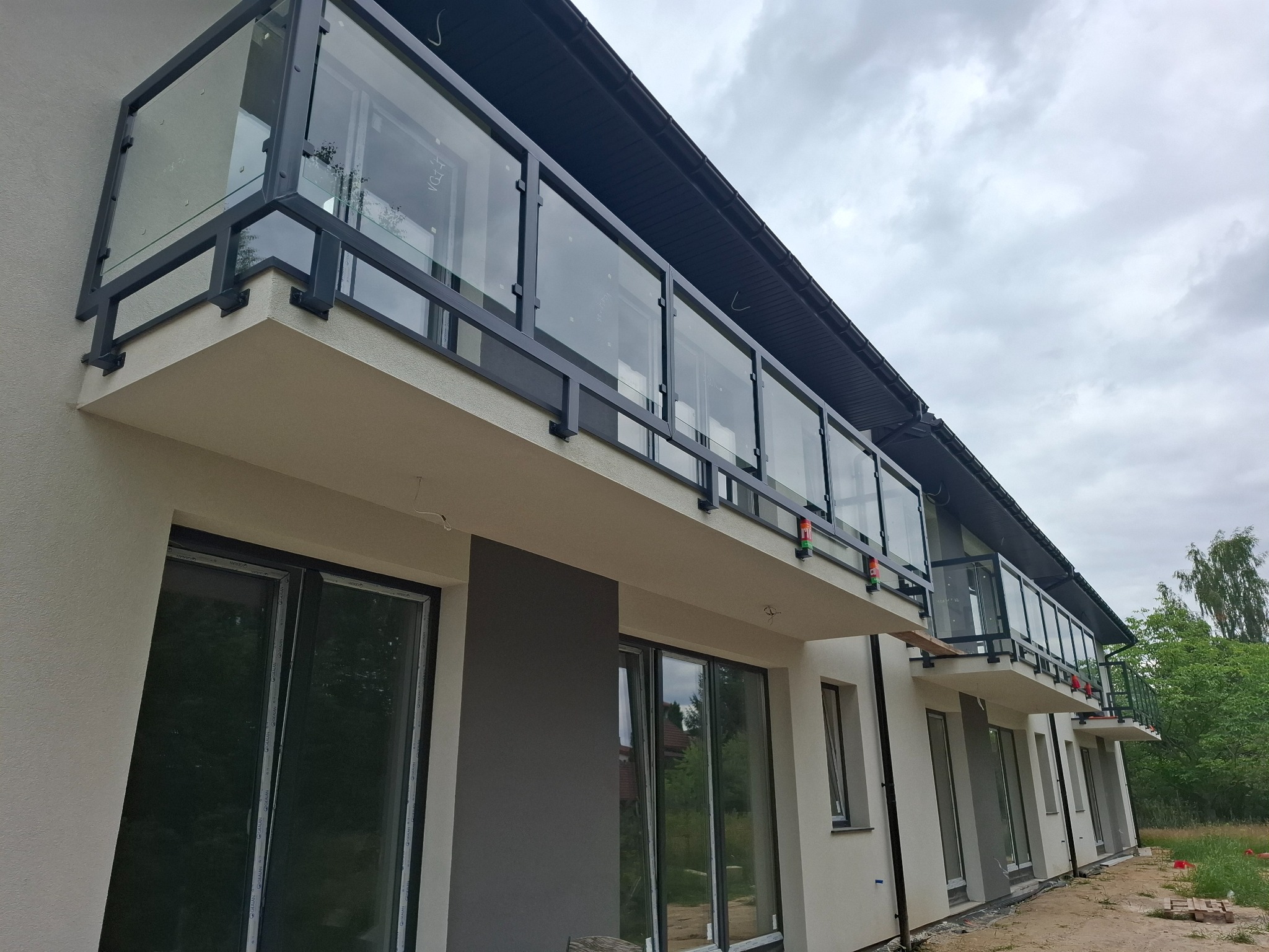 Szereg balkonów z czarnymi metalowymi ramami i szklanymi balustradami w nowym budynku mieszkalnym, widok z dołu na pochmurne niebo.