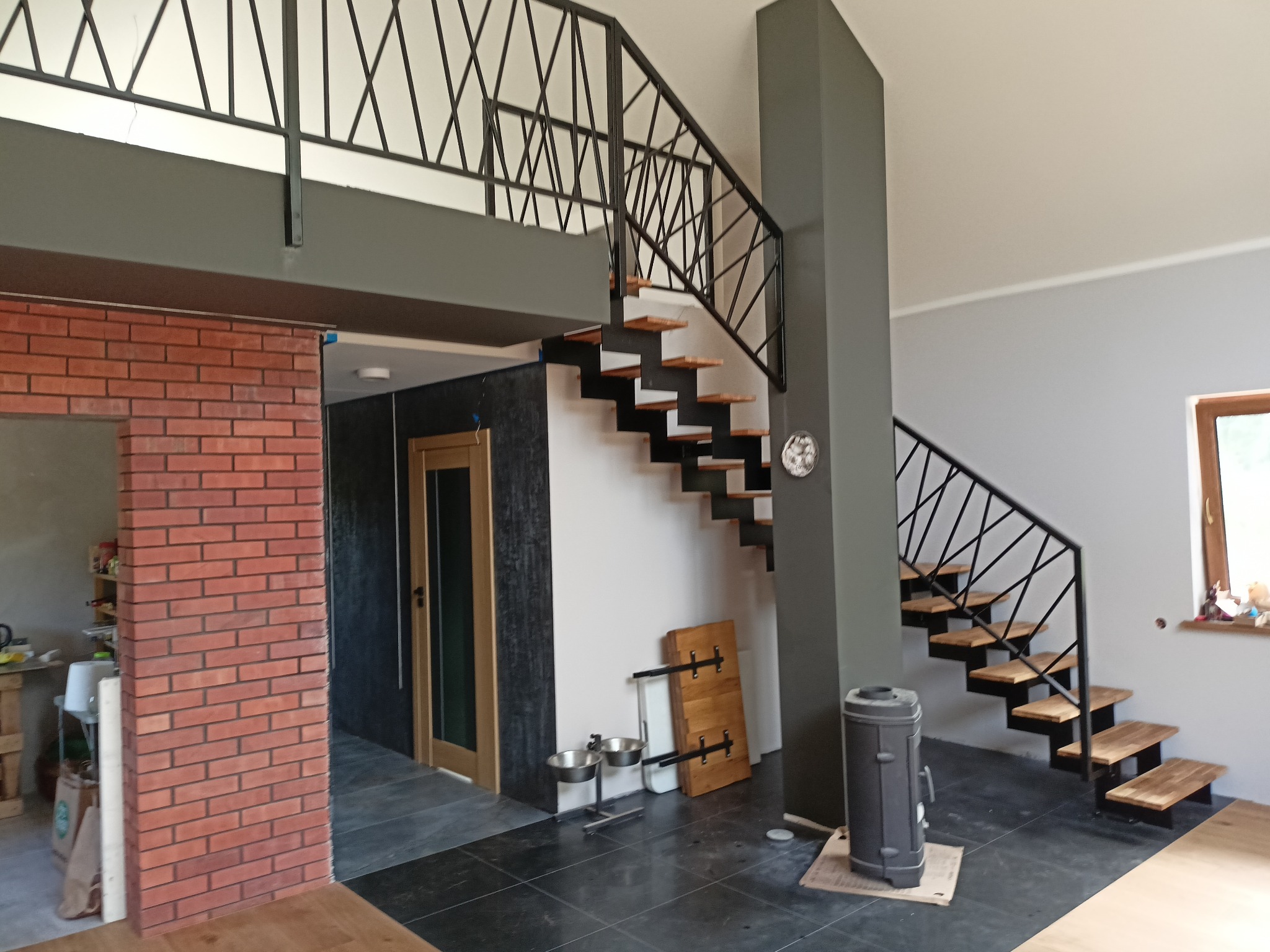 Nowoczesne dwubiegowe schody z drewnianymi stopniami i czarną metalową balustradą o geometrycznym wzorze, widoczne w przestronnym wnętrzu z ceglaną ścianą i szarą kolumną.