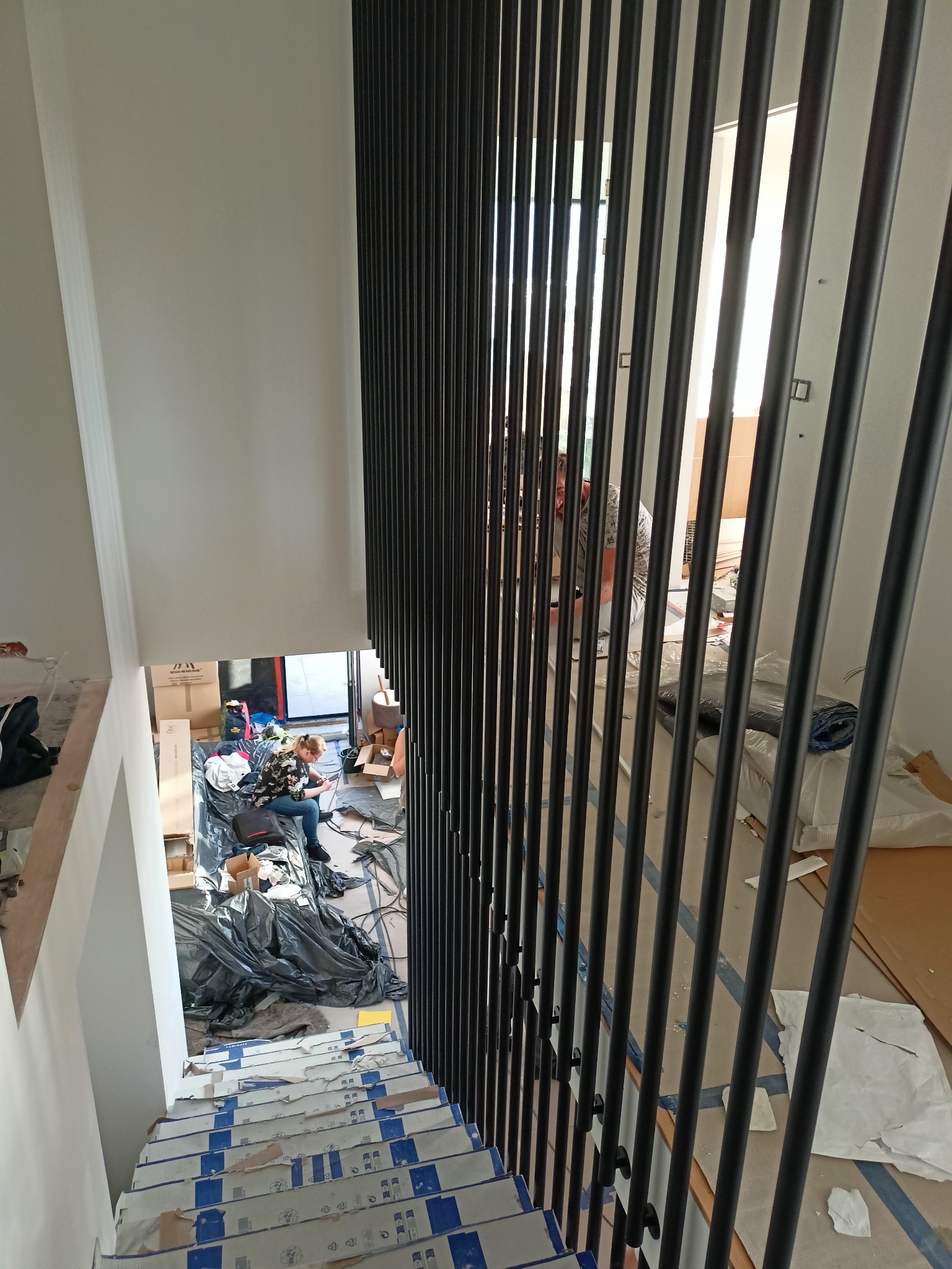 Wysoki kąt widzenia na montaż balustrady metalowej ze szczeblami w nowoczesnym domu w trakcie wykończenia, z widocznymi zabezpieczonymi schodami i osobą pracującą na dole.