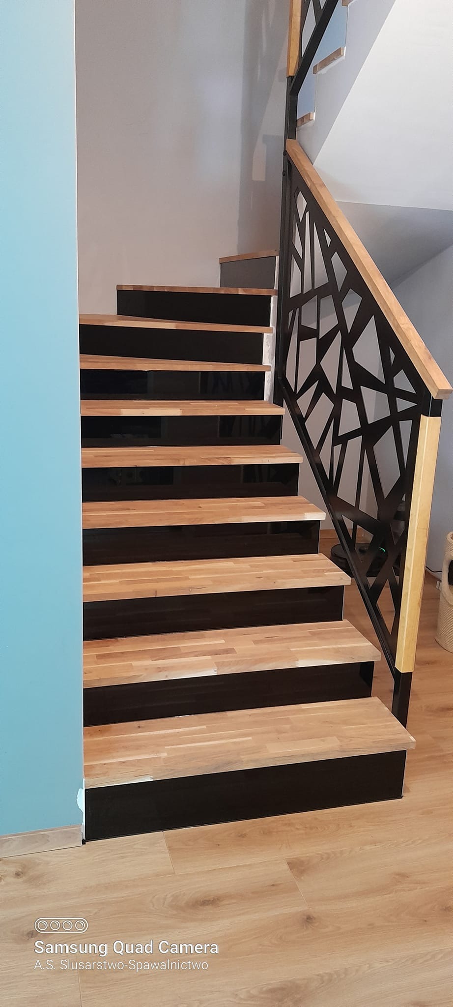 Nowoczesne schody z drewnianymi stopniami i czarnymi podstopnicami, z metalową balustradą o geometrycznym wzorze i drewnianym poręczą.