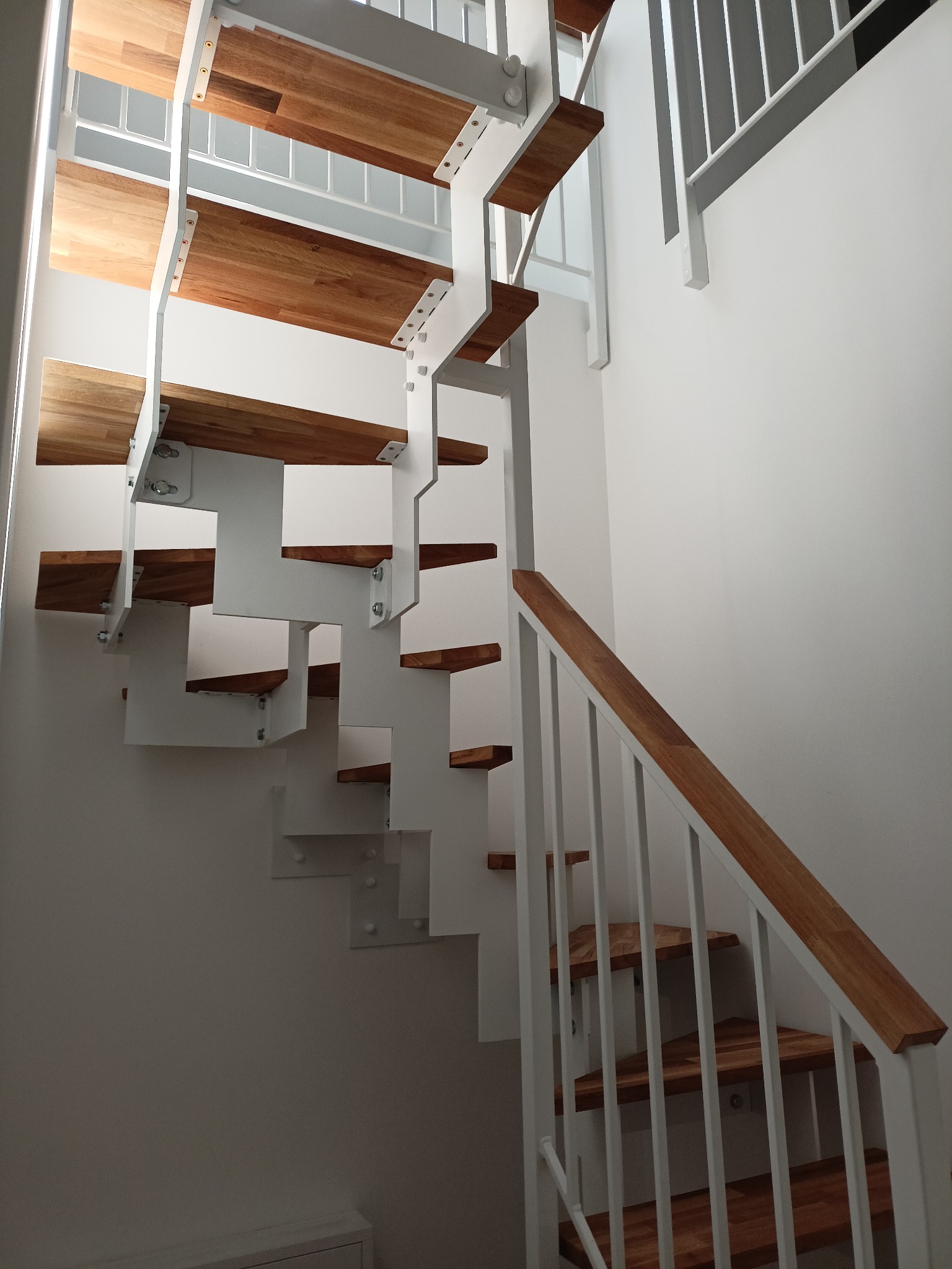 Nowoczesne schody z drewnianymi stopniami i białą metalową konstrukcją, mocowane śrubami, z białą balustradą, w jasnym wnętrzu.