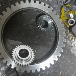 Zestaw trzech kół zębatych o różnej wielkości, ułożonych na brudnym, betonowym podłożu w warsztacie, obok leżą narzędzia i pojemnik z opiłkami.