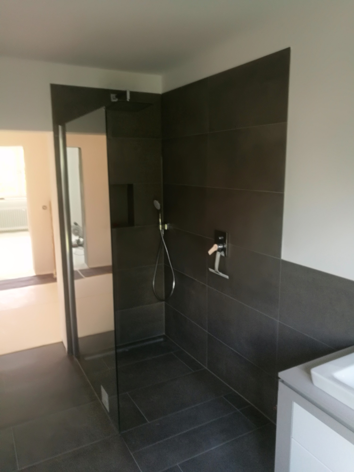 Nowoczesna kabina prysznicowa typu walk-in z ciemnymi płytkami, deszczownicą i wnęką na kosmetyki, widok na sąsiednie pomieszczenie.