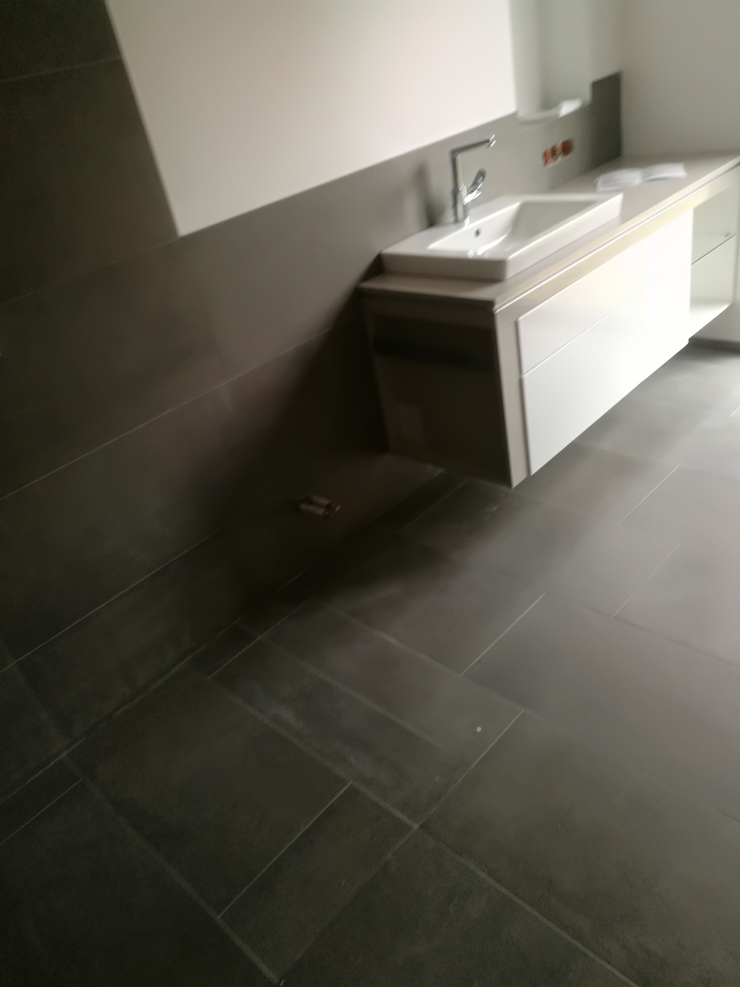 Nowoczesna umywalka z szafką w łazience, szare płytki podłogowe ułożone w prostokąty, minimalistyczny design, bateria chromowana.