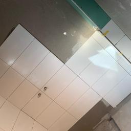 Pobrzut - Układanie białych, błyszczących płytek na ścianie, widoczne krzyżyki dystansowe i taśma maskująca, surowy tynk na ścianie i fragment płyty kartonowo-gipsowej.