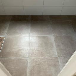 Pobrzut - Beżowe płytki podłogowe i białe płytki ścienne w łazience, widoczne z góry przez otwarte drzwi. Część podłogi zabezpieczona kartonem z napisem 'fiora'.