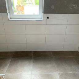 Pobrzut - Ściana i podłoga wyłożone jasnymi płytkami, widoczne okno z białymi ramami oraz surowy betonowy sufit w trakcie prac wykończeniowych.