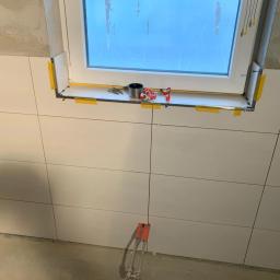 Pobrzut - Białe płytki glazurnicze ułożone na ścianie wokół okna, z widocznymi narzędziami glazurniczymi na parapecie i wystającymi rurami instalacji wodnej w dolnej części ściany.