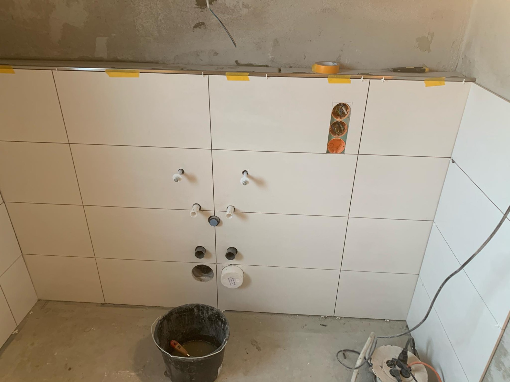 Ściana w trakcie układania białych, kwadratowych płytek, widoczne otwory na instalacje, taśma malarska zabezpieczająca górną krawędź, czarne wiadro z narzędziami na podłodze.