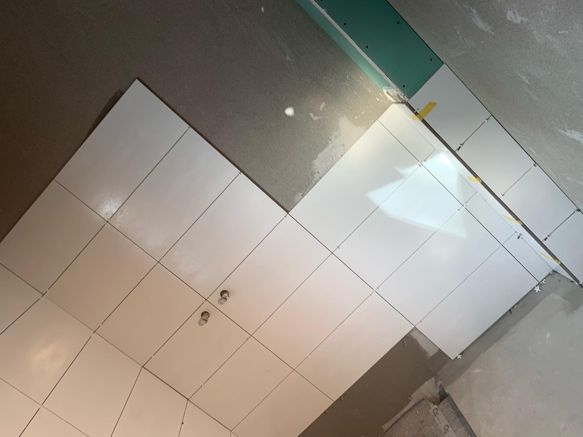 Układanie białych, błyszczących płytek na ścianie, widoczne krzyżyki dystansowe i taśma maskująca, surowy tynk na ścianie i fragment płyty kartonowo-gipsowej.