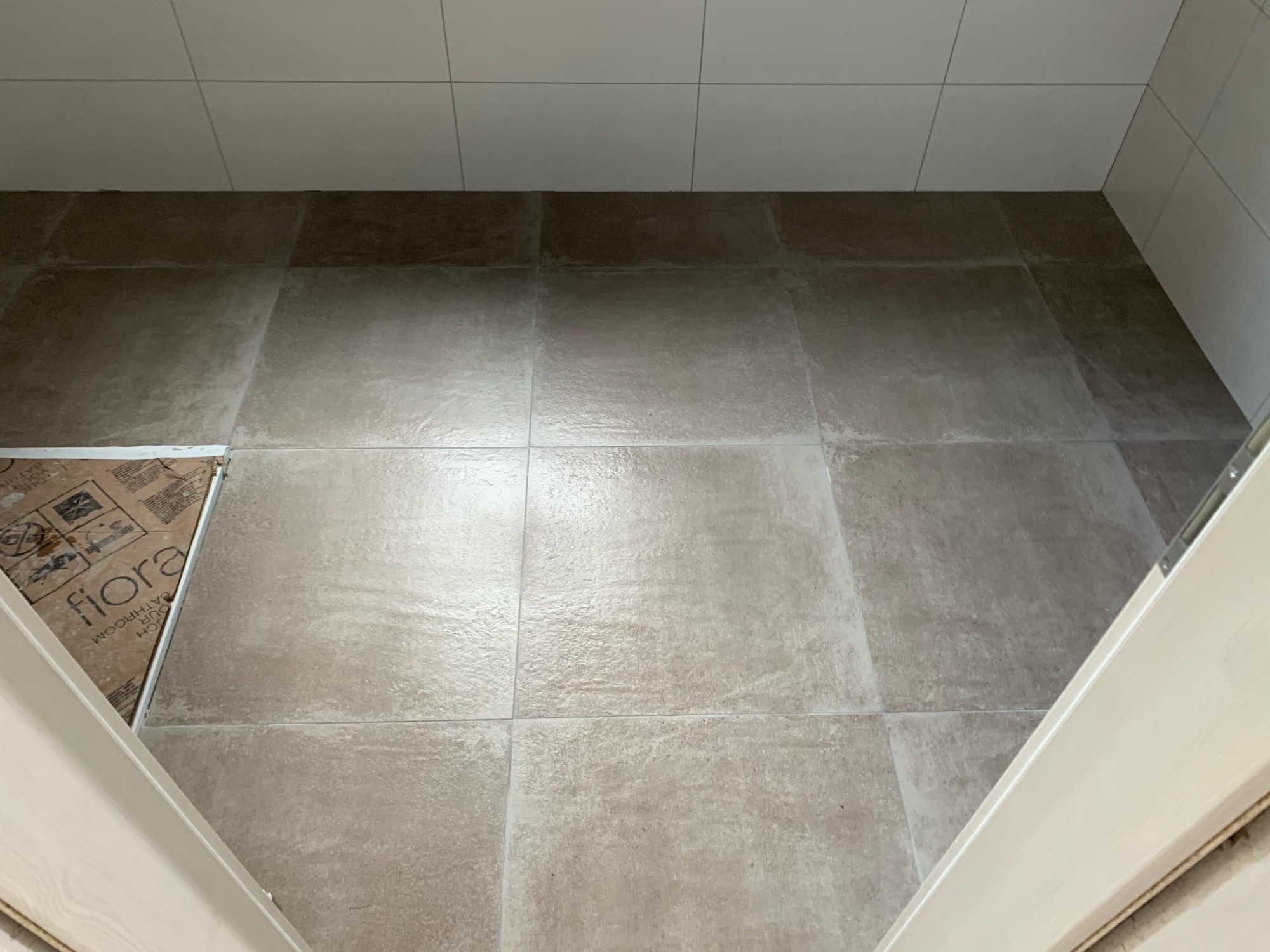 Beżowe płytki podłogowe i białe płytki ścienne w łazience, widoczne z góry przez otwarte drzwi. Część podłogi zabezpieczona kartonem z napisem 'fiora'.