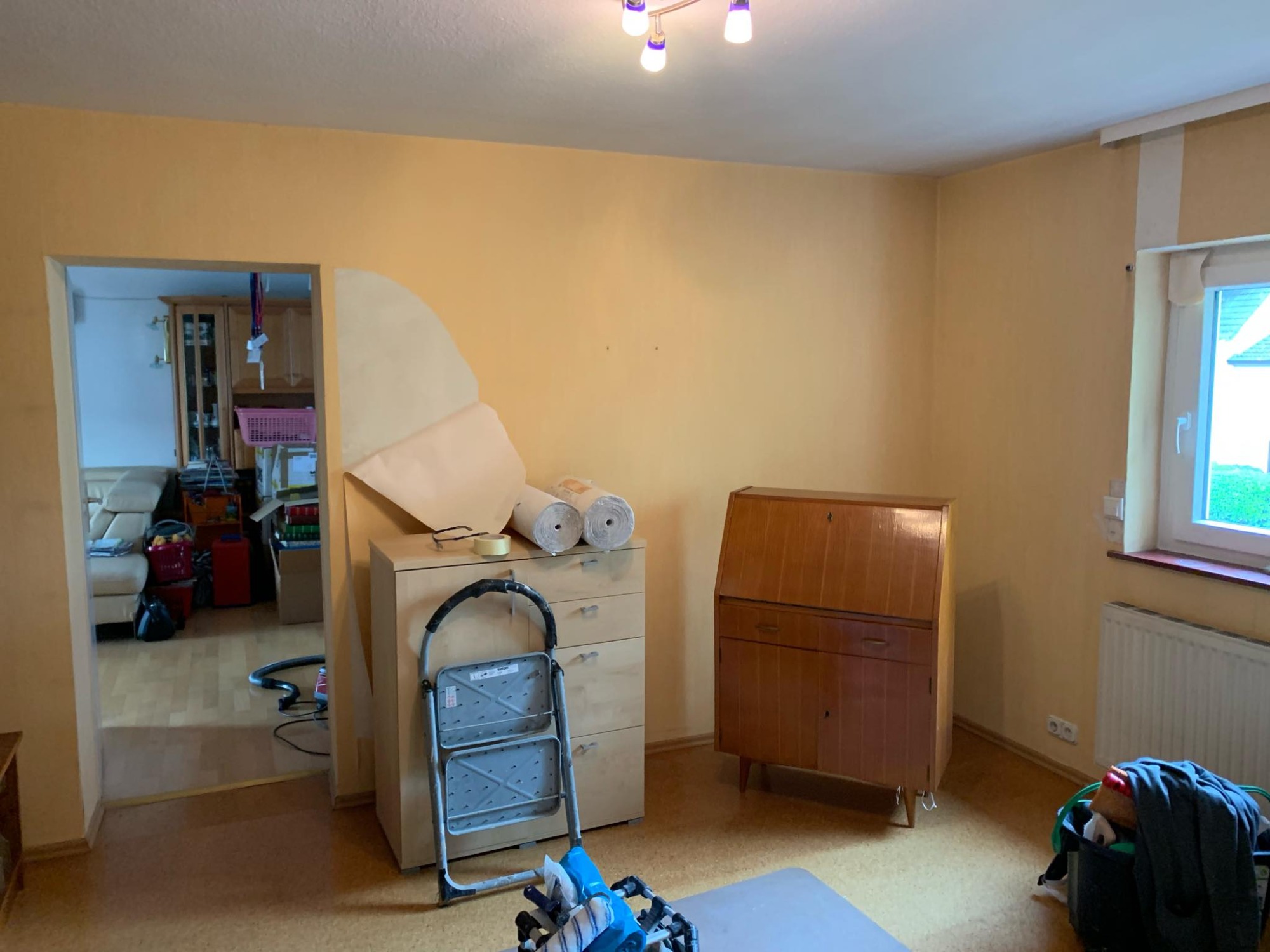 Pokój w trakcie przygotowań do malowania: częściowo oklejona ściana, drabina, wałki malarskie, meble odsunięte od ścian.