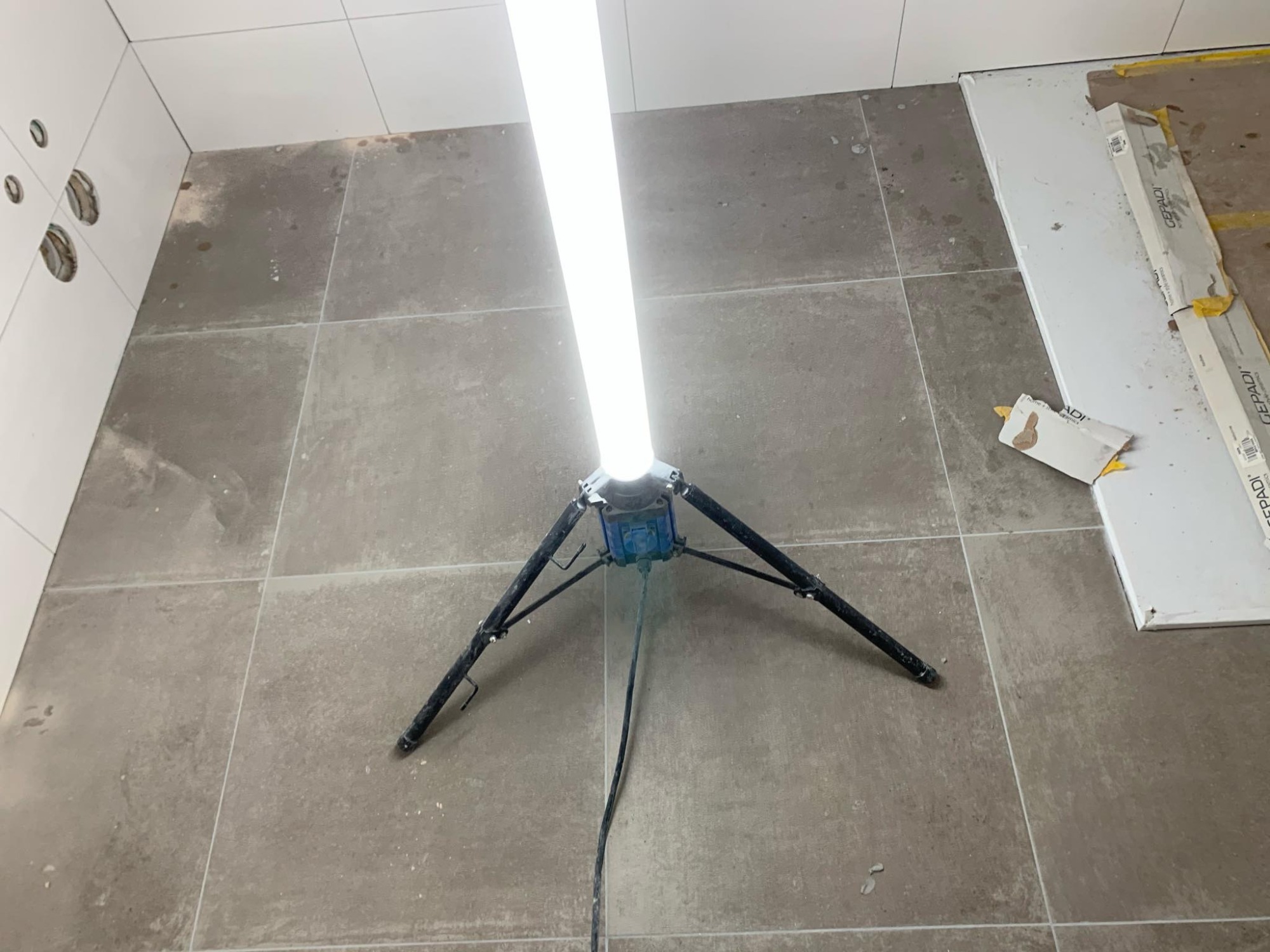 Podłoga w trakcie układania szarych płytek, oświetlona lampą budowlaną na trójnogu, widoczne białe płytki na ścianie i opakowania po płytkach pod ścianą.