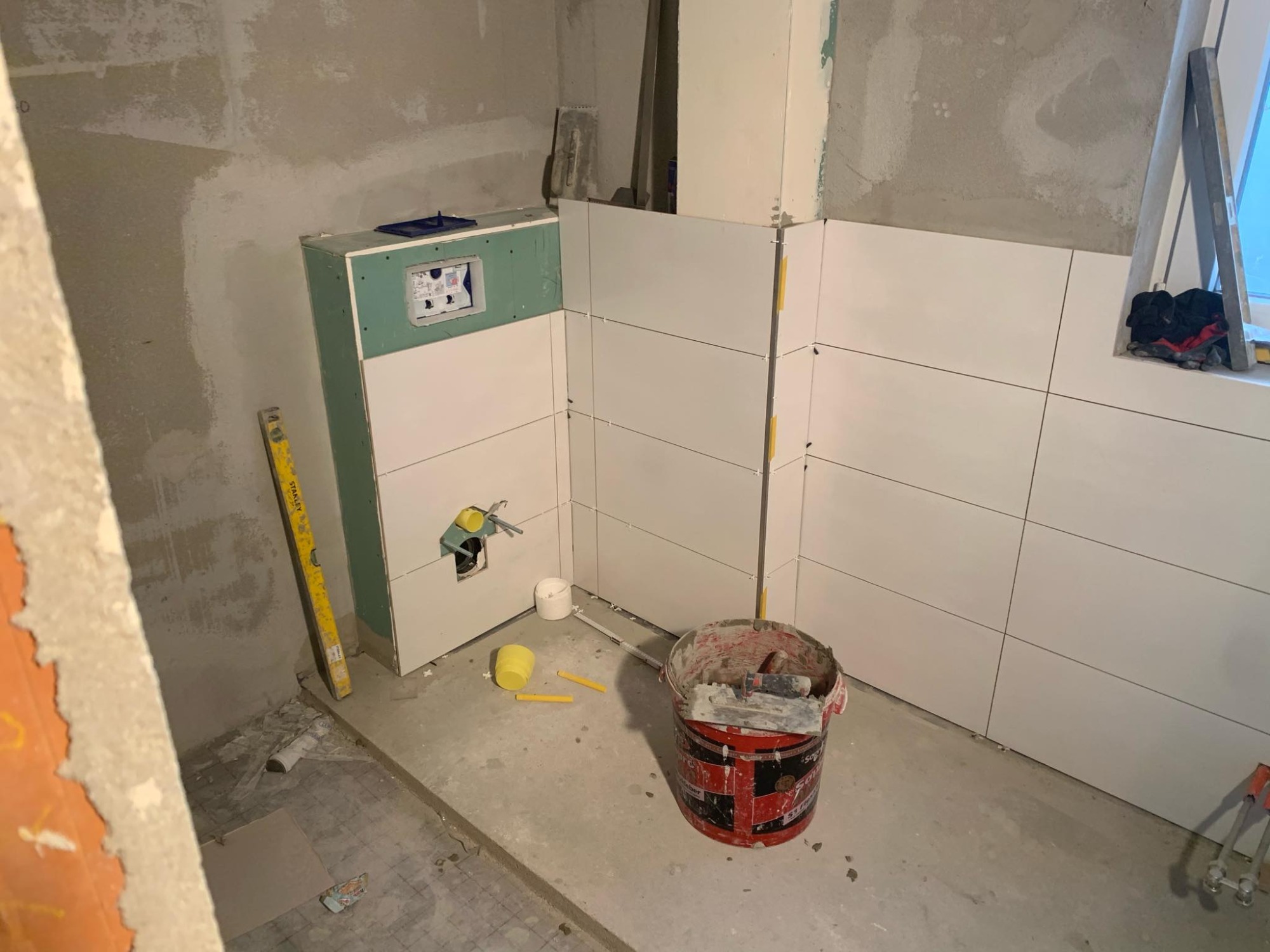 Białe płytki ułożone na ścianie w łazience w trakcie remontu, widoczny stelaż podtynkowy WC obudowany płytą gipsowo-kartonową, poziomica Stanley i wiadro z narzędziami glazurniczymi.