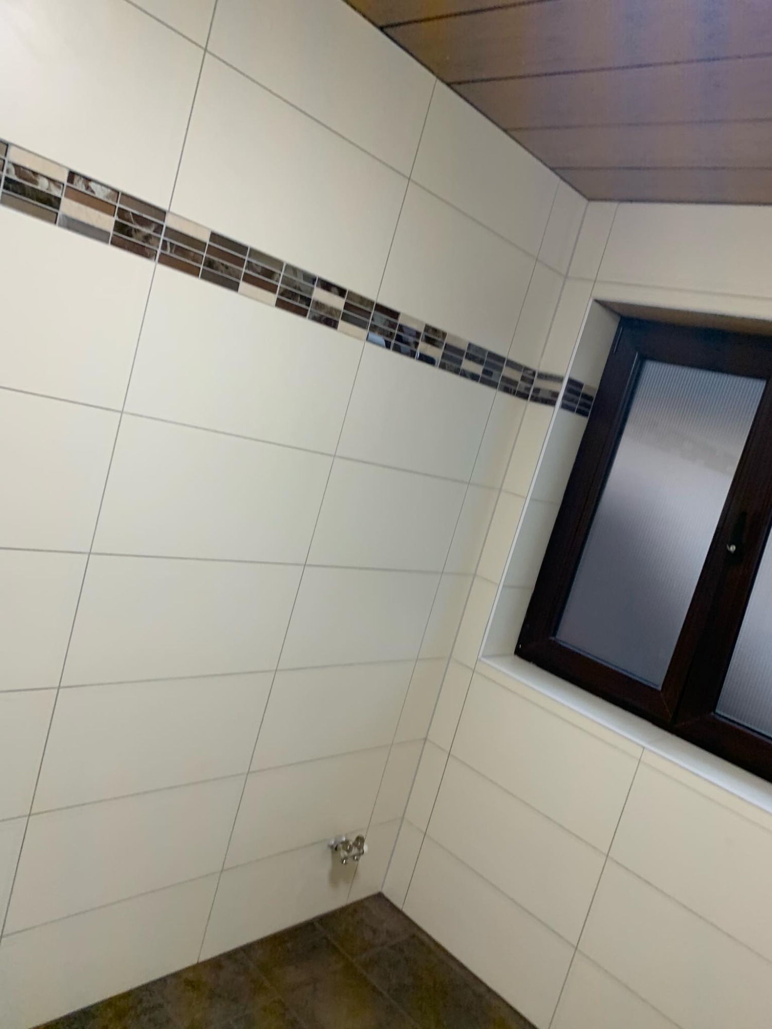 Wykończona łazienka z białymi, kwadratowymi płytkami, brązowym pasem dekoracyjnym i brązową ramą okna z żaluzjami wewnątrz szyby.