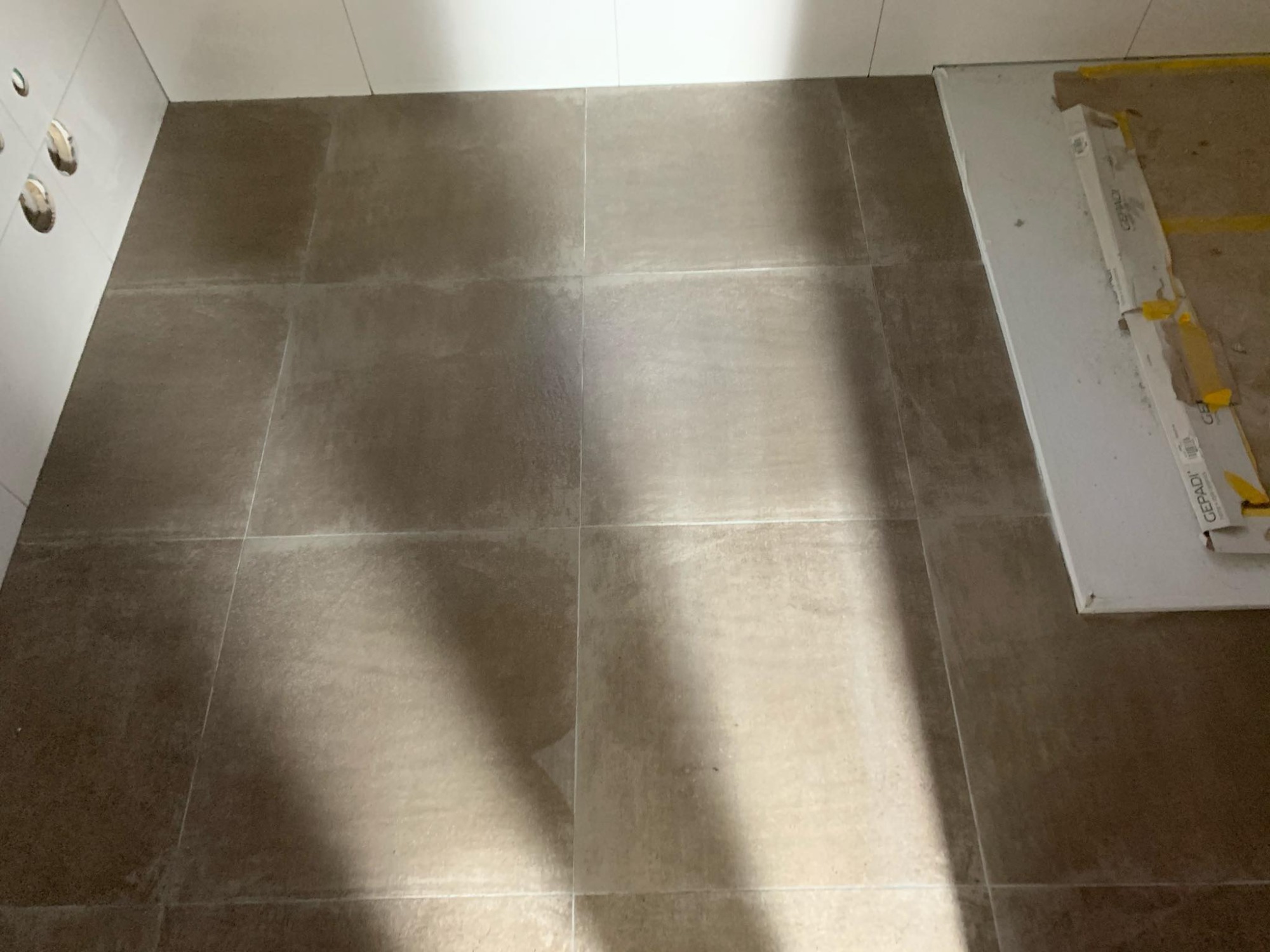 Ułożona podłoga z płytek ceramicznych w odcieniach szarości, widoczne białe ściany z otworami na instalacje oraz resztki opakowania po płytkach przy ścianie.