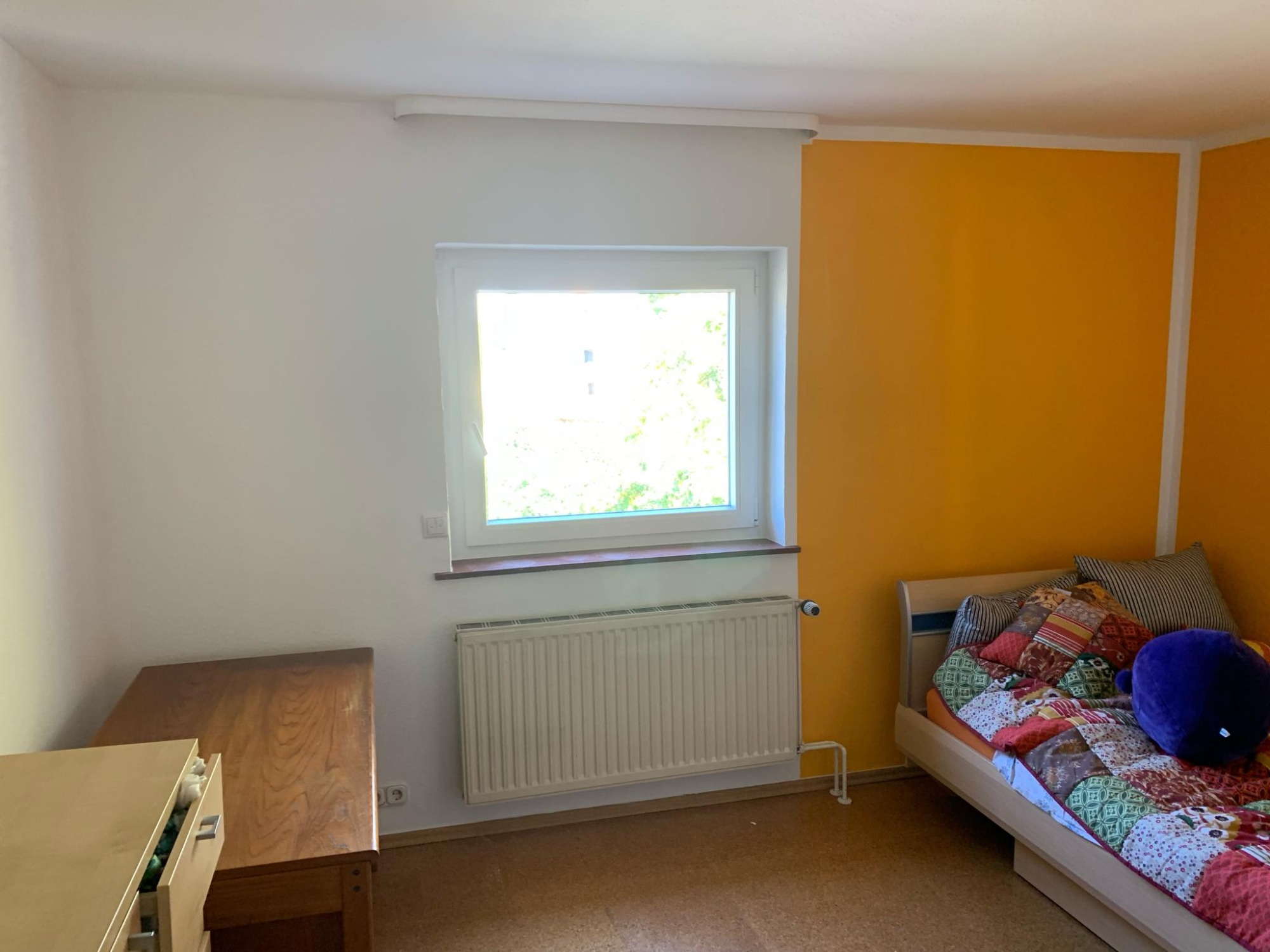 Jasny pokój z pomarańczową ścianą, białym grzejnikiem pod oknem i patchworkową narzutą na łóżku
