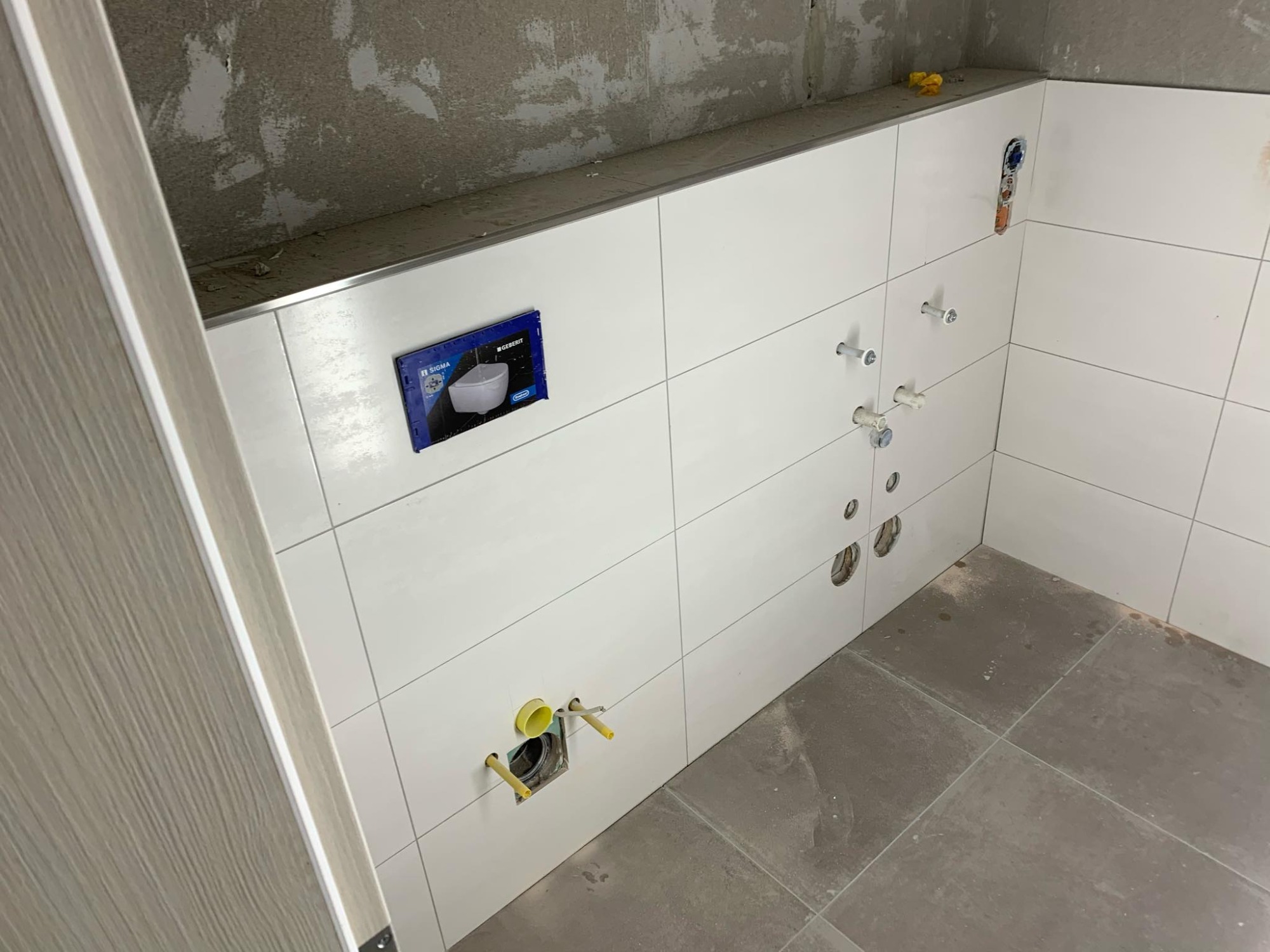 Białe płytki ścienne w trakcie montażu w łazience, widoczne otwory na instalacje, szara podłoga, oraz częściowo wykończona ściana nad płytkami.