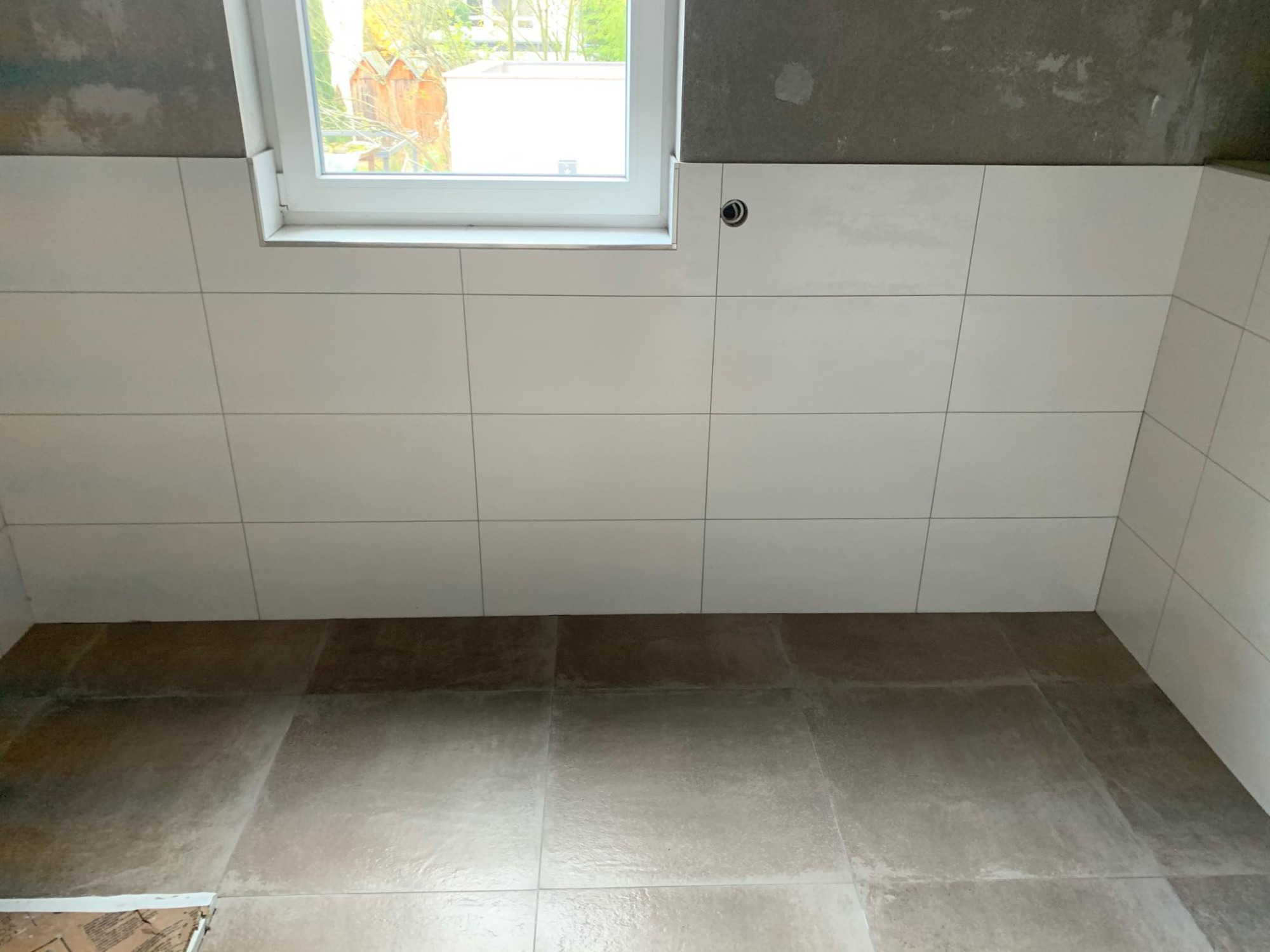 Ściana i podłoga wyłożone jasnymi płytkami, widoczne okno z białymi ramami oraz surowy betonowy sufit w trakcie prac wykończeniowych.