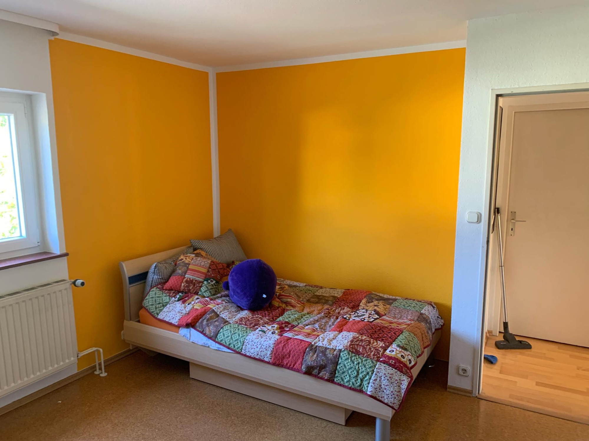 Pokój z pomalowanymi na żółto ścianami, łóżkiem z patchworkową narzutą i fioletową poduszką, widoczny grzejnik i odkurzacz w przejściu do innego pomieszczenia.