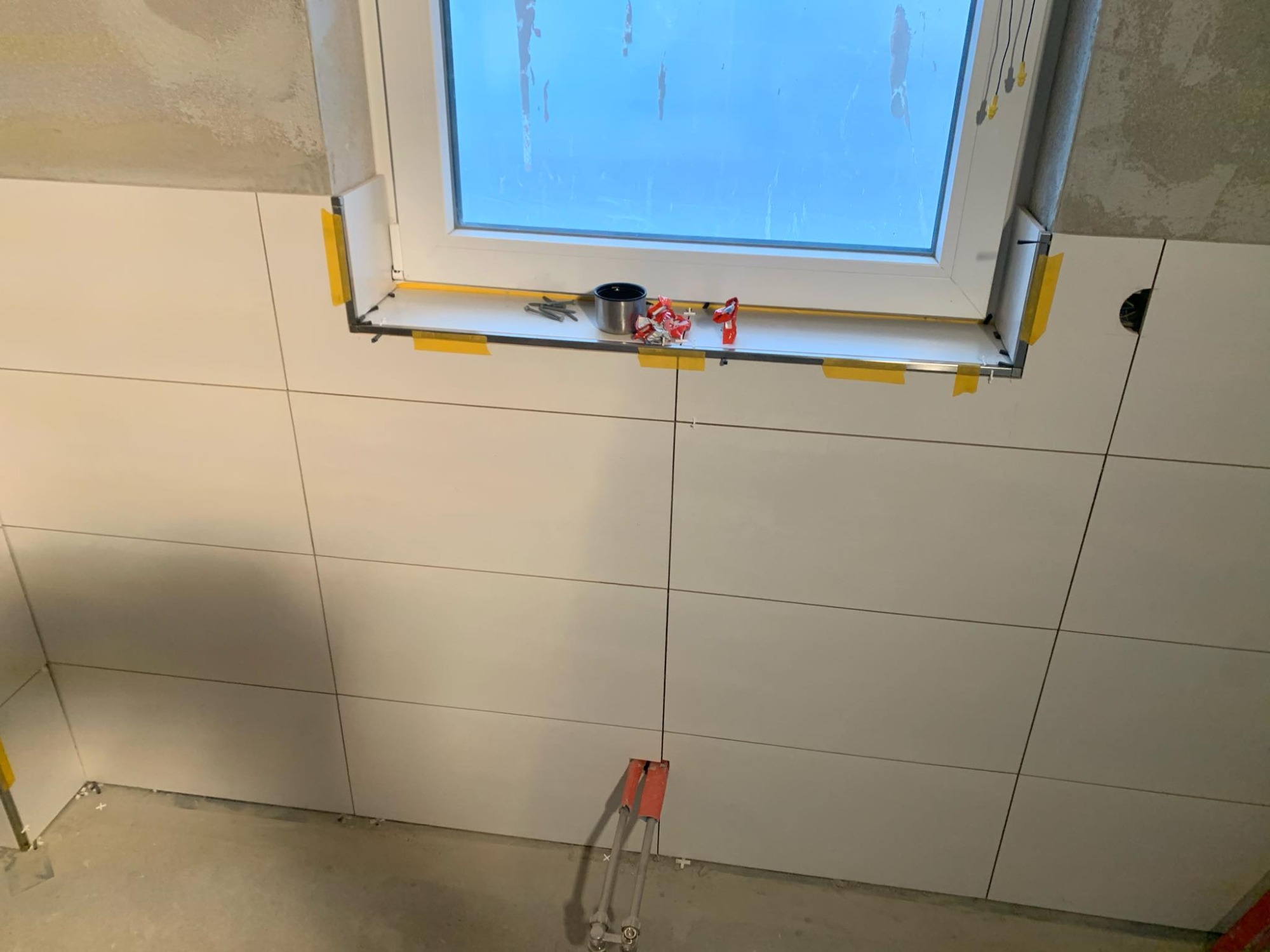 Białe płytki glazurnicze ułożone na ścianie wokół okna, z widocznymi narzędziami glazurniczymi na parapecie i wystającymi rurami instalacji wodnej w dolnej części ściany.
